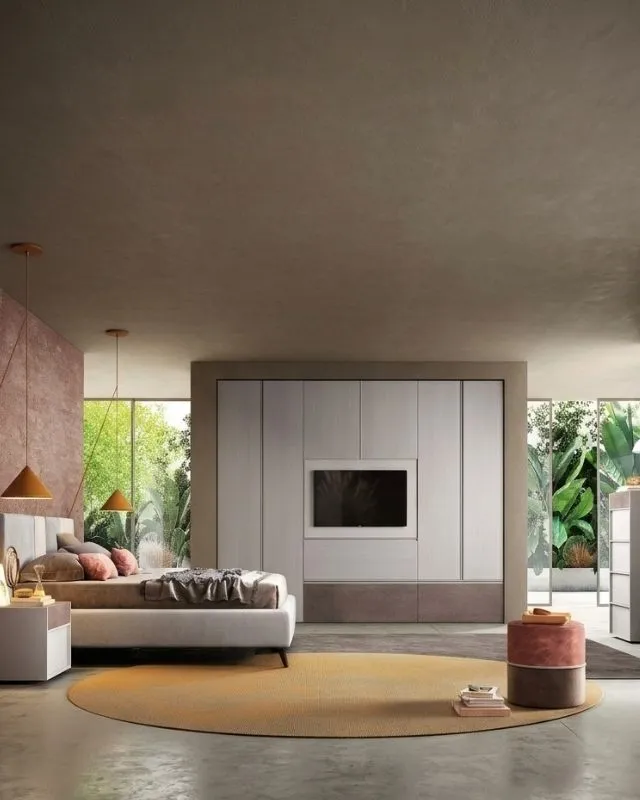 Dettaglio verticale di un'accogliente camera da letto Behome. L'inquadratura si concentra sulla zona notte, con un letto matrimoniale finemente allestito. Sulla destra, è visibile la sezione centrale del capiente armadio a muro chiaro con modulo dedicato alla televisione.