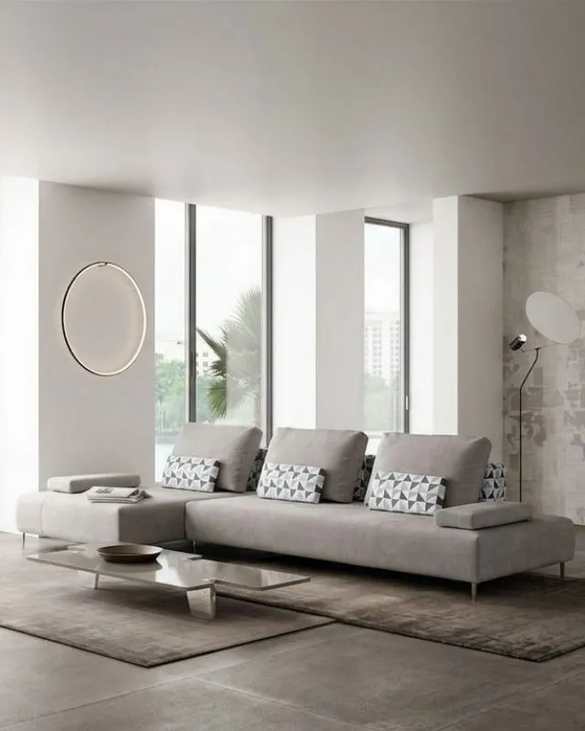 Dettaglio di un moderno divano angolare grigio chiaro Behome con cuscini geometrici, inserito in un luminoso soggiorno con tavolino basso e una lampada circolare a parete.
