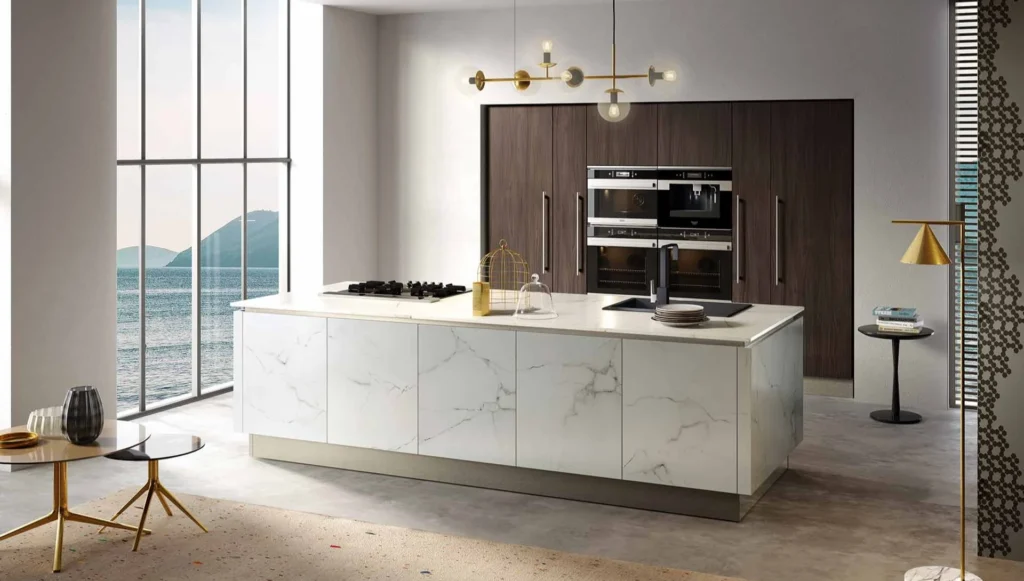 Ampia Cucina Moderna Dafne Even con isola in marmo bianco, colonne in legno scuro, forni a incasso e vista mare.