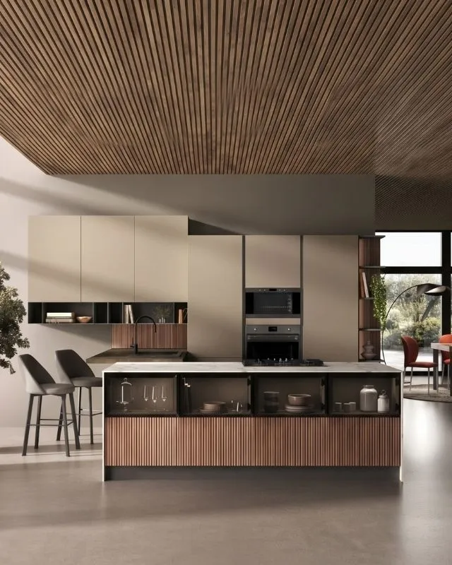 Dettaglio verticale di una cucina dal design moderno Behome. L'inquadratura si concentra sull'isola centrale, caratterizzata da un top in marmo chiaro, piano cottura e una base con eleganti vetrinette espositive posizionate sopra un rivestimento cannettato.
