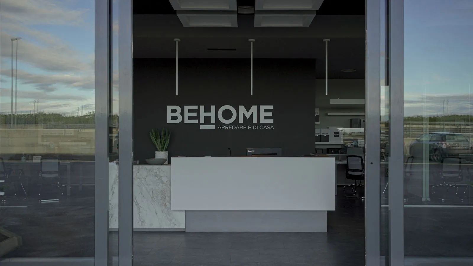 Ingresso principale della sede Behome con porte scorrevoli a vetri aperte. All'interno si intravede una reception moderna con bancone bianco e marmo, sormontata da una grande parete nera con il logo aziendale bianco "BEHOME - ARREDARE È DI CASA"